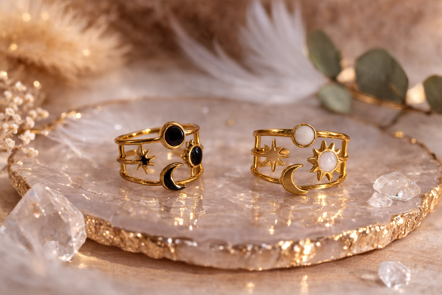 Bague ajustable plaquée or avec motif soleil et lune, ornée d’une pierre noire ou blanche, posée sur une coupelle en pierre avec décor naturel et plumes en arrière-plan