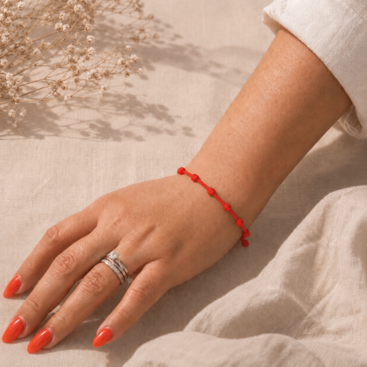 Main portant un bracelet rouge à sept nœuds sur fond en lin beige, avec une lumière douce et des fleurs séchées.