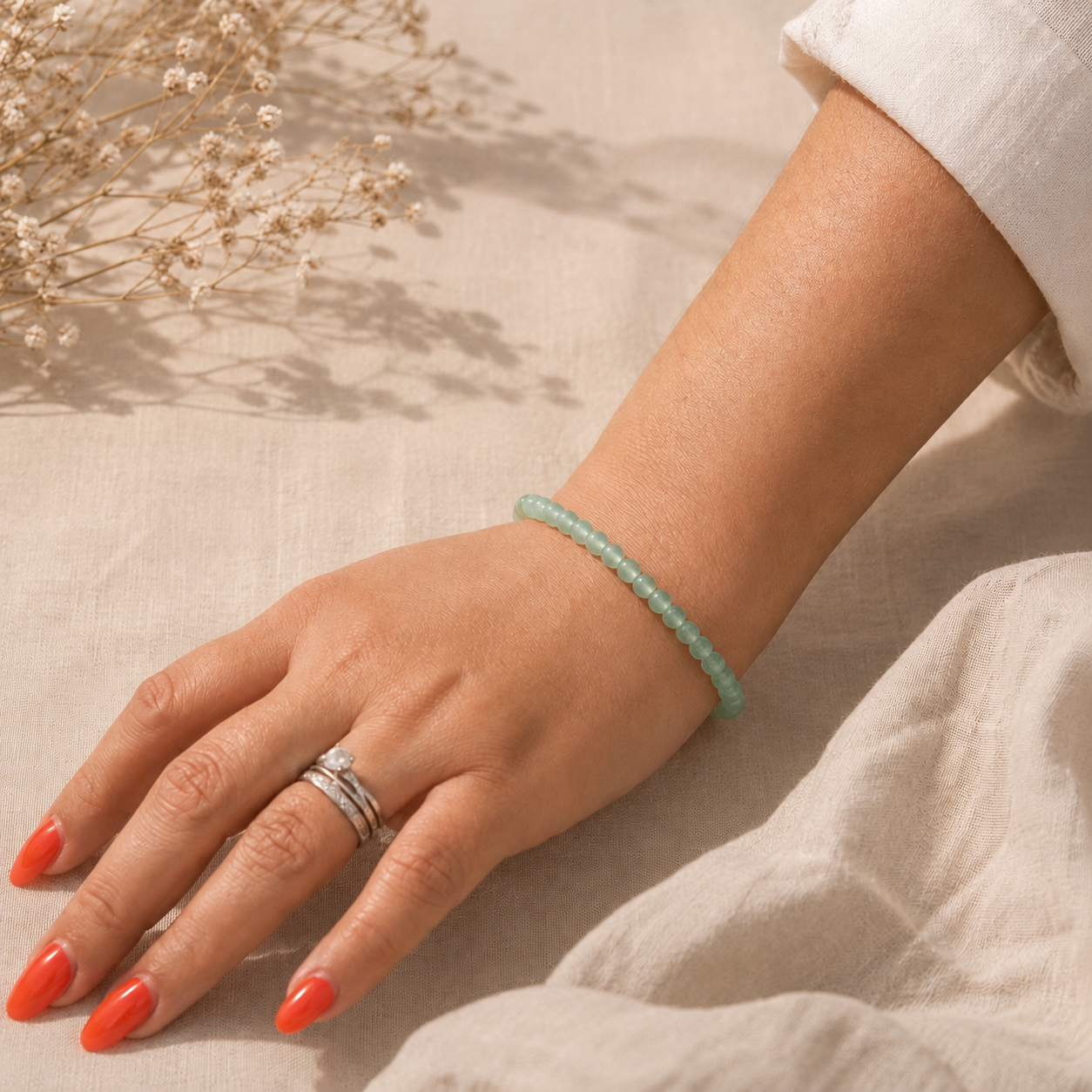 Main portant un bracelet fin en aventurine verte naturelle, posé sur un fond en lin beige avec une lumière douce et des fleurs séchées.