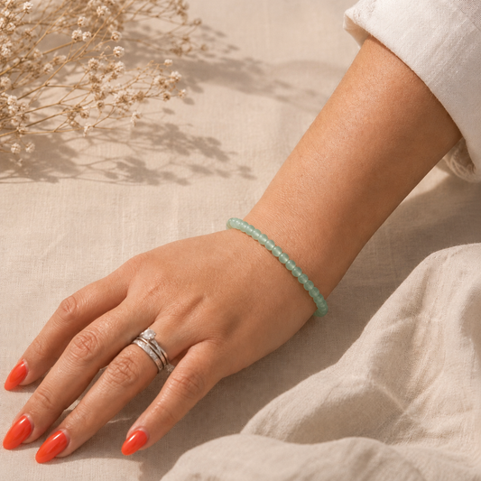 Main portant un bracelet fin en aventurine verte naturelle, posé sur un fond en lin beige avec une lumière douce et des fleurs séchées.