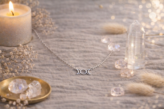 Collier triple lune en acier inoxydable posé sur un tissu beige avec bougie, cristaux de quartz et fleurs séchées dans un décor doux et spirituel