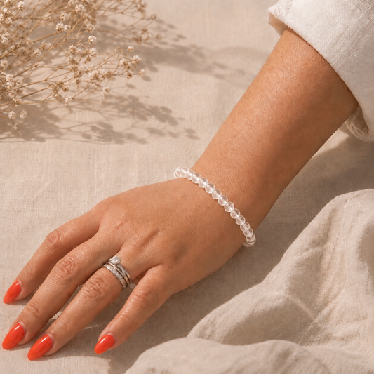 Main portant un bracelet en Cristal de Roche naturel sur fond en lin beige, avec une lumière douce et une tenue claire.