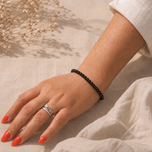 Main portant un bracelet fin en obsidienne noire naturelle sur fond en lin beige, avec une lumière douce, une tenue claire et des fleurs séchées.