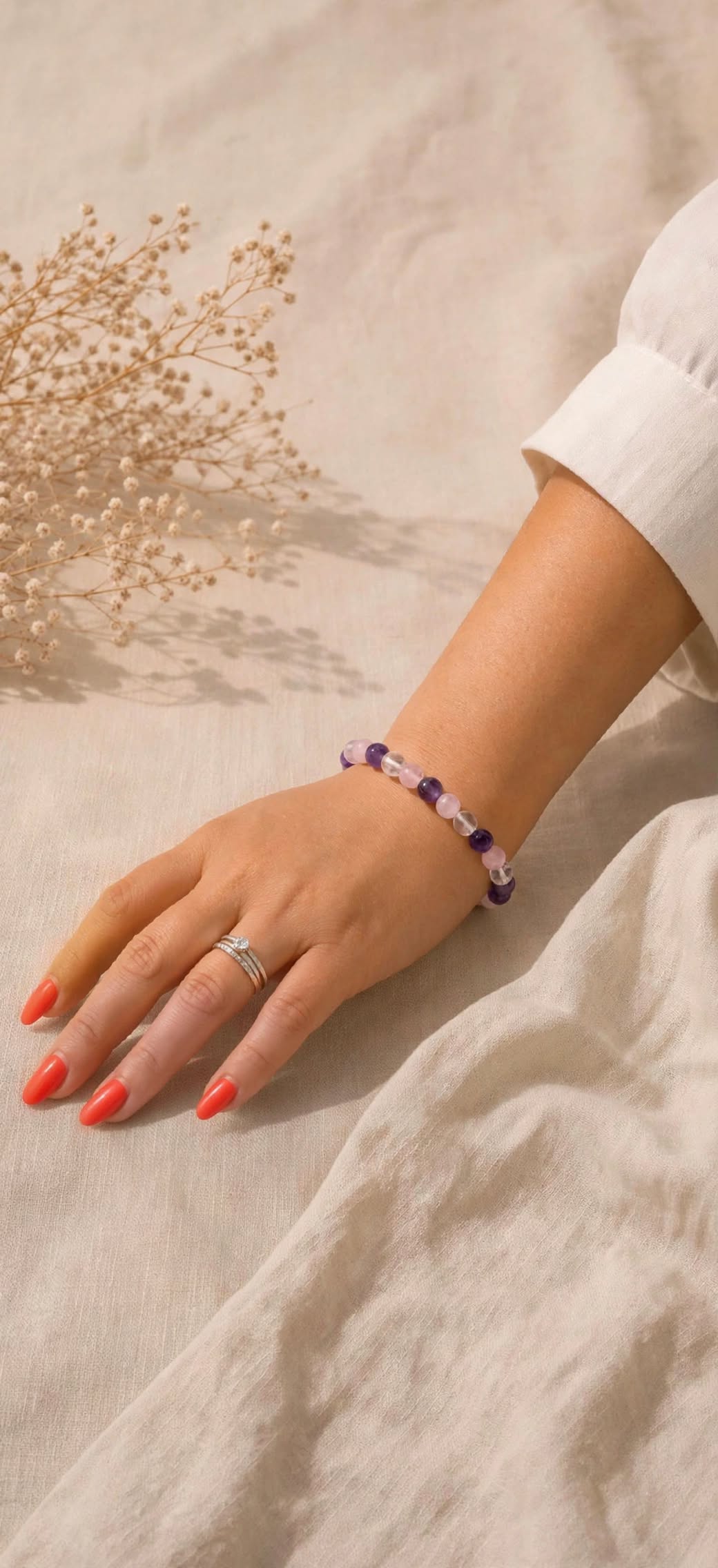 Bracelet Harmonie en améthyste violette, quartz rose et cristal de roche porté au poignet d'une femme, vernis corail, décor lin beige et gypsophile séché
