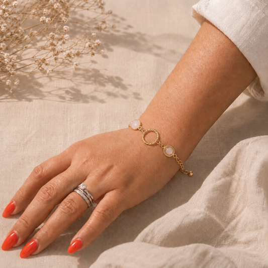 Main portant un bracelet doré avec pierres en quartz rose et anneau torsadé, sur fond en lin beige et lumière douce.