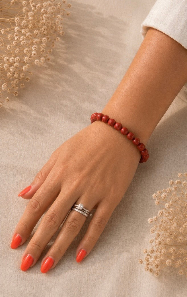 
Main portant un bracelet en jaspe rouge naturel sur fond en lin beige, avec une lumière douce, une tenue claire et des fleurs séchées.