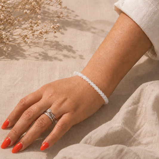 Main portant un bracelet fin en Pierre de Lune naturelle sur fond en lin beige, avec une lumière douce, une manche claire et des fleurs séchées.