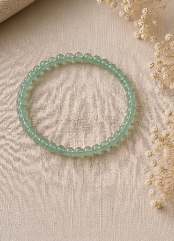 un bracelet fin en aventurine verte naturelle, posé sur un fond en lin beige avec une lumière douce et des fleurs séchées.